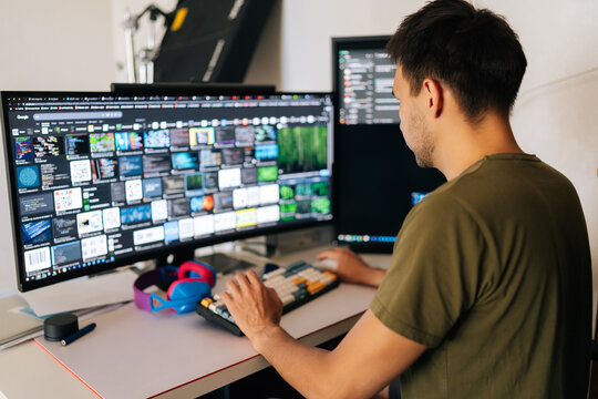 Back view of software developer male writing code at home office desk, surrounded by dual monitors displaying lines of code and communication platforms in modern workspace. Concept of remote workplace