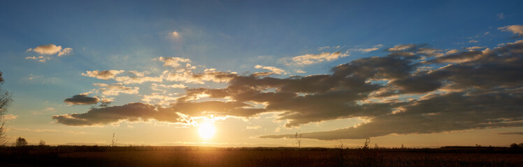 Fototapeta premium Sun sets low on horizon, casting golden glow across landscape. Fluffy clouds, illuminated with warm hues, float against clear blue sky. Serene scene tranquil, with last light of day fading gently.
