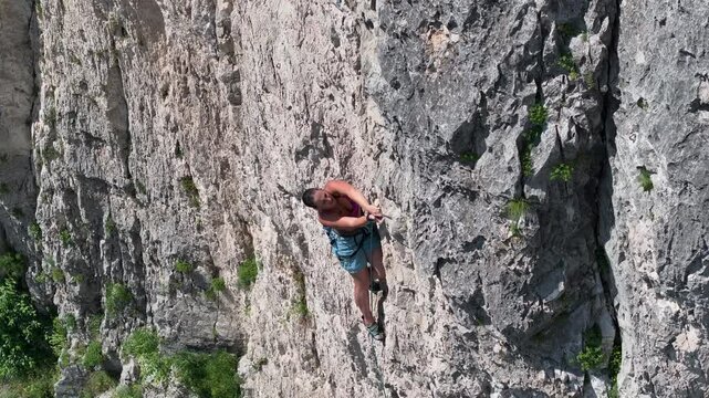 AERIAL: Woman in the middle of climbing on a rugged, vertical rock face. With her gaze fixed on the route, she reaches into chalk bag and continues. Challenging and exciting of the outdoor sport.