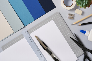 White sheet with ruler and knife on cutting mat among craft tools and colored paper. Creative workspace for art and design preparation.