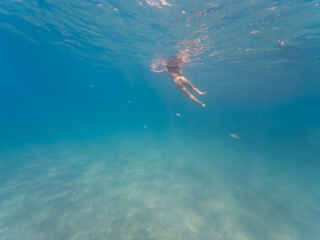 A serene underwater scene shows a woman in a bikini swimming in the crystal-clear turquoise sea on a sunny day.