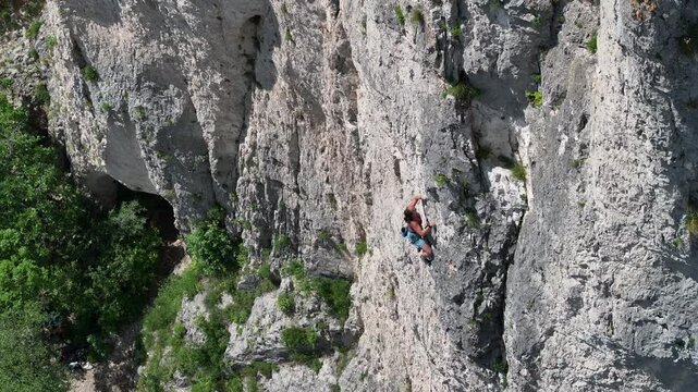 AERIAL: Female rock climber lead climbing a steep rock face on a sunny day. Scenic vertical wall is surrounded by lush greenery. Challenging adrenaline sports activity in beautiful natural environment