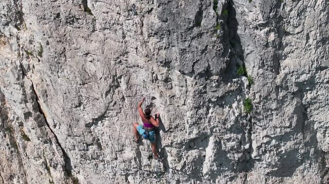 AERIAL: Brave woman in her climbing gear searches for good grip, as she navigates a difficult section of a rock wall. Her focus and body position illustrate the skill and physical demands of the climb