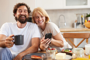 Couple sits at a wooden table in a bright kitchen, enjoying breakfast with coffee and food. They are smiling and looking at a phone, sharing a joyful moment together.