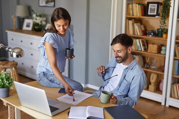 Obraz premium A woman leans over a desk to point at notes while a man sits casually, holding a mug and smiling. Sunlight fills the home office, creating a warm and inviting atmosphere.