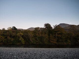Sundown and moon rising on the Nevis Range mountains above the River Lochy and the autumnal woodland on it's bank, seen from Camaghael,  Highlands of Scotland.