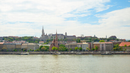 The stunning skyline of Budapest, Hungary, showcasing its historic buildings along the riverbank. Trees offer greenery against the backdrop of distant towers and landmarks.