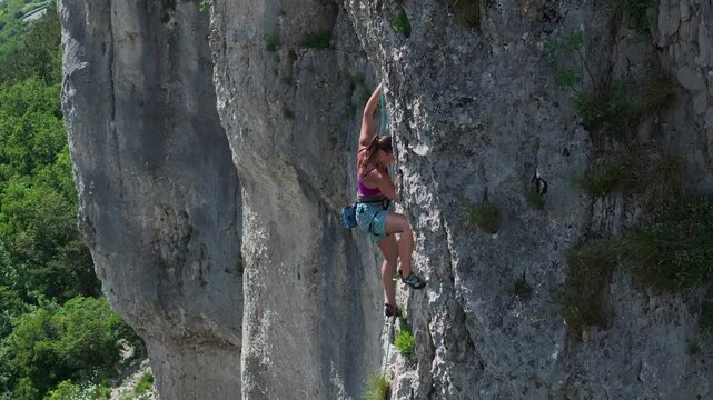 AERIAL: Female athlete is in the midst of a climb, reaching for a handhold on rocky wall. Equipped with harness and secured by rope, she is lead climbing a challenging route in limestone rock face.