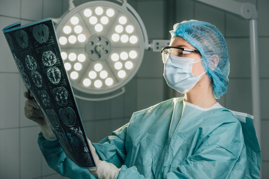 Surgeon in green scrubs and protective gear, examining medical imaging in an operating room, illuminated by bright surgical lights, showcasing dedication to patient care and precision in healthcare