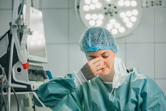 A female surgeon in a green gown and surgical cap looks tense as she sits in an operating room surrounded by medical equipment and bright surgical lighting.
