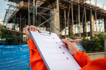 Action of an engineer is checkling on safety checklist form at construction working site (with background of house builing). Industrial safe work practice concept, close-up with selective focus.