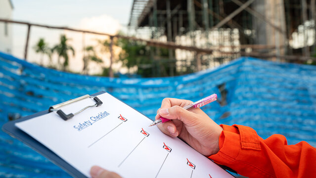 Action of an engineer is checkling on safety checklist form at construction working site (with background of house builing). Industrial safe work practice concept, close-up with selective focus.