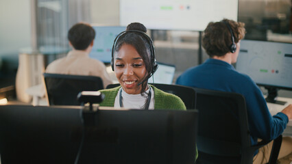Smiling customer service representative wearing headset works at computer in modern office, with two colleagues in background also engaged in similar tasks