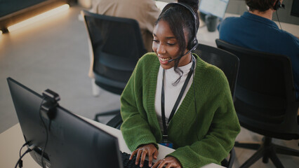 Smiling customer service representative wearing green sweater and headset works at computer in...