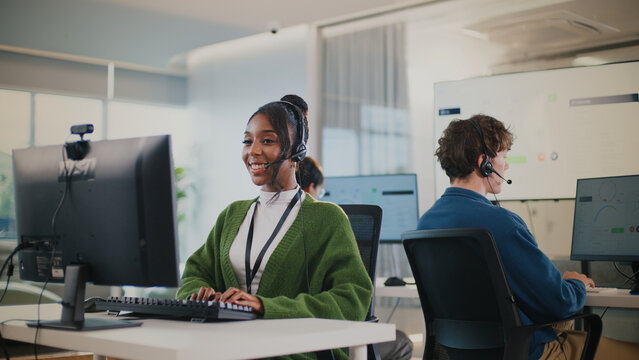 Cheerful customer service representative wearing headset is working at computer in modern office environment. Other team members are visible in background, collaborating in professional setting