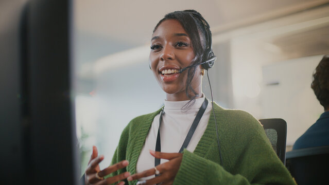 Cheerful customer service representative wearing headset and green sweater is engaging in conversation while seated at her workstation in modern office environment