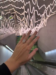 Vertical close up photo of a woman's hand wearing a large oval cut diamond engagement ring, held up against a modern escalator with artistic neon lights in the background.