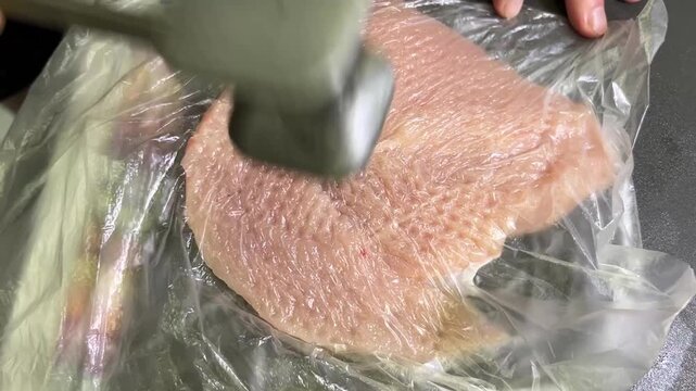 Raw chicken breast covered with plastic wrap being pounded flat with a meat mallet for cooking