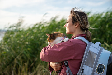 Content happy smiling woman walking hiking carrying cat enjoying sunshine grassy field. Pet owner with backpack carrier for pets traveling, outdoor adventure, wildlife cat care, feline companion walk
