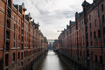 Naklejka premium Historical warehouse district in Hamburg features red brick buildings along a scenic canal under a cloudy sky