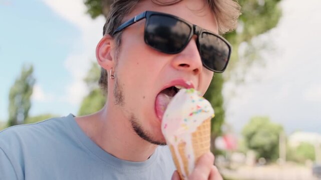 Young man in sunglasses licking pastel ice cream cone in a close-up summer shot. Focus on mouth, tongue, and melting dessert with natural outdoor light and greenery in the background.