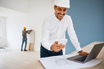 Smiling Architect Using Mobile Phone With Laptop and Blueprints on Construction Site