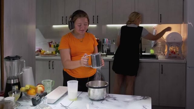 A cheerful 11-year-old girl in headphones smiles and bops her head to music while sifting flour into a bowl in modern kitchen, mother is washing dishes in background. Family moment of joyful cooking.