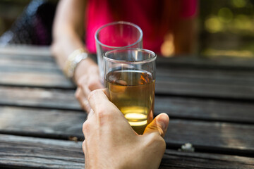 inquadratura con punto di vista e da vicino di due persone adulte, che fanno un brindisi con due bicchieri pieni di varie bevande, sedute ad un tavolo di legno, all'aperto, di giorno