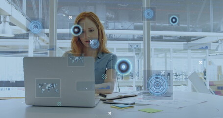 Woman in light blue shirt typing on laptop in office, with notepads and data graphics