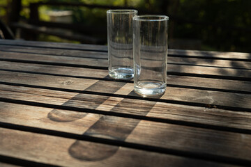 bicchieri di vetro, trasparenti e vuoti, su un tavolo di legno, illuminati dal sole, che proiettano le loro ombre lunghe sulla superficie