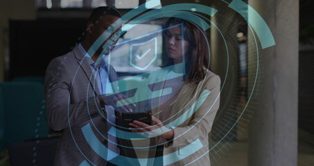 Two colleagues wearing suits reviewing tablet in office corridor, with holographic security overlay