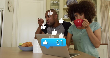 Mid adult couple eating breakfast while using silver laptop at wooden kitchen table, with red mug