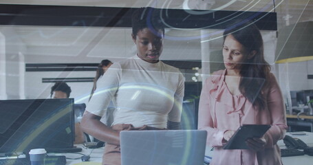 Collaborating women wearing office attire using overlays on laptop, tablet in office, with monitors