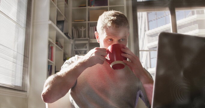 Sipping coffee man checking laptop screen at home office, with red mug, blinds and bookshelf
