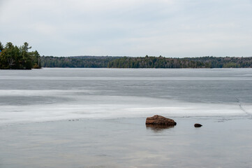 A peaceful horizontal landscape of a large, partially frozen lake with a lone rock protruding from the smooth ice, with a forest on the far shore.