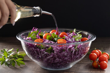 A hand is pouring a liquid, likely oil or dressing, from a clear glass bottle into a large clear glass bowl filled with a fresh 