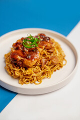 Side angle view of fried noodles topped with grilled chicken served on white plate with blue background, Asian food presentation concept.
