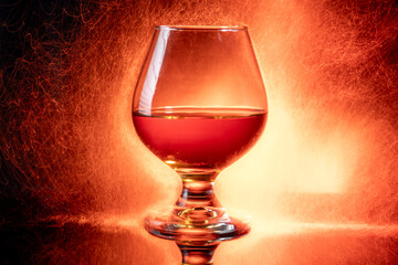 A snifter glass filled with a reddish-brown alcoholic beverage, likely brandy or cognac, set against a warm, fiery