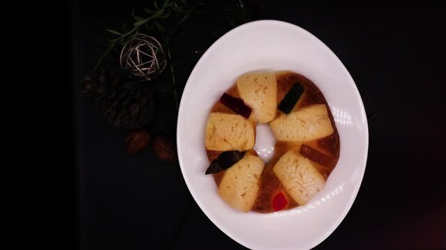 Traditional Rosca de Reyes bread in Mexico on white plate  black background turning alongside the walnut and fresh rosemary decorations lock dawn camera top view