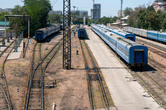 Railway station yard with blue carriages on parallel tracks, semaphore signal, concrete platforms, overhead line supports, turnout switches. Passenger service layover, marshalling activities