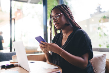 Concentrated young Black woman checking phone while working on laptop, immersed in digital tasks, reflecting flexibility and multitasking in mobile tech-based freelance environment.