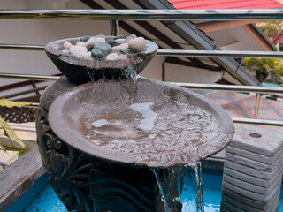 A close-up view of a tiered garden fountain with water flowing from a stone bowl filled with smooth rocks, set against a blurred background.