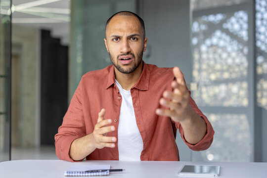 A man in casual attire talking and gesturing passionately, looking towards the camera. The setting is contemporary with a clean design and a notepad and phone visible on the table. - Powered by Adobe
