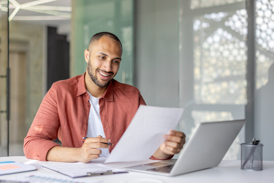 A cheerful individual analyzes paperwork at their desk while working on a laptop in a sleek, contemporary office environment. The scene conveys professionalism, productivity, and relaxed confidence - Powered by Adobe