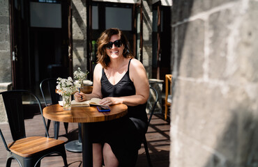 Smiling mature woman in sunglasses writing in notebook while using smartphone, combining analog reflection with digital efficiency in smart, modern lifestyle.