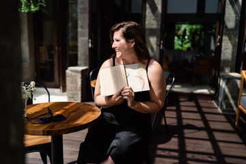 Joyful woman laughing and looking away while showing open sketchbook, enjoying creative freedom, relaxed mood and nature-inspired workspace with smartphone set aside.
