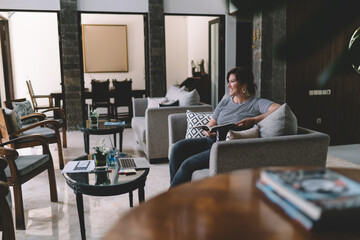 Fototapeta premium Freelancer smiling with book in hand, seated comfortably beside laptop, enjoying mindful moment of calm in digitally driven, coworking inspired interior.
