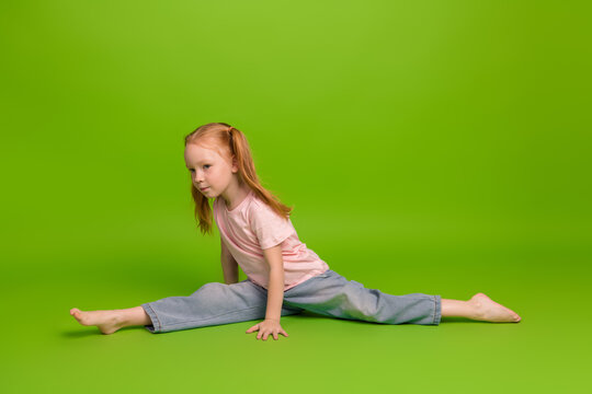 Cute girl displaying flexibility by performing a split on a bright green background wearing casual attire