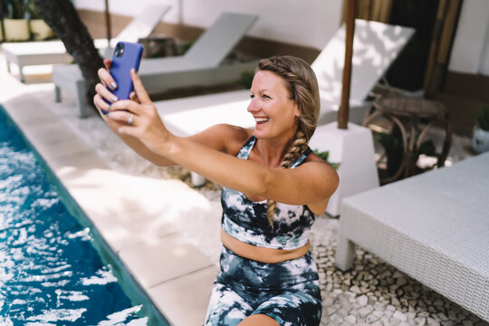 Smiling blonde woman in sportswear takes selfie by pool, enjoying carefree moment of digital connection. Self-love, confidence, and healthy lifestyle meet modern tech in joyful setting.
