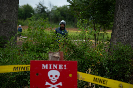 A serious photojournalistic image capturing a red minefield warning sign with a skull symbol, highlighted by yellow hazard tape in an outdoor field setting.

